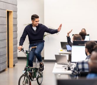 Young man riding a bicycle in office and waving hand to colleagues. Hispanic man on cycle greeting colleagues in a coworking office.